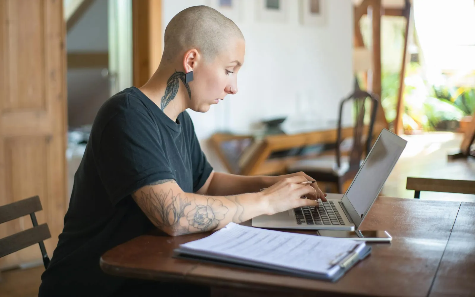 Pessoa trabalhando em um notebook à mesa de uma casa iluminada, com uma prancheta de papéis à frente